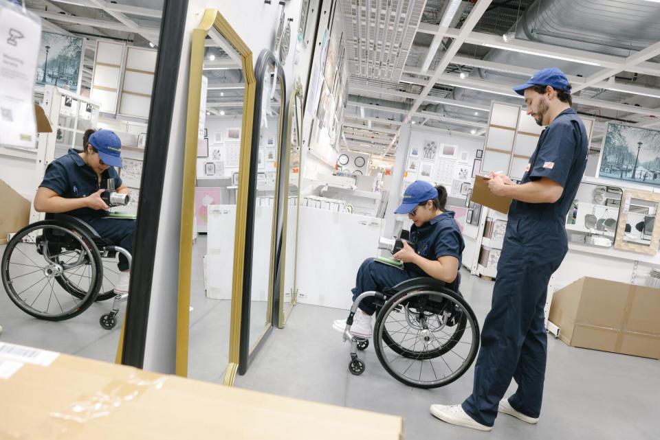 A woman in a wheelchair photographs herself in front of a floor-length mirror. A man stands next to her.