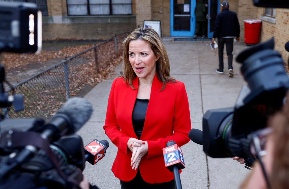 Jocelyn Benson, in red jacket, speaks in front of a barrage of media microphones.