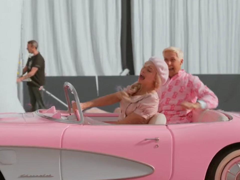 Barbie actors Ryan Gosling and Margot Robbie in pink car in front of men in black with cameras