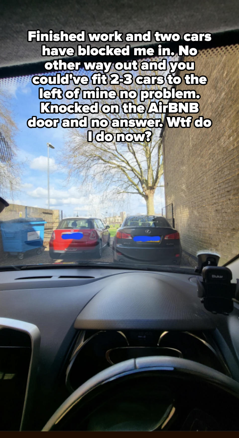 View from a car's driver seat showing two parked cars ahead on a narrow street with a tree and clear sky