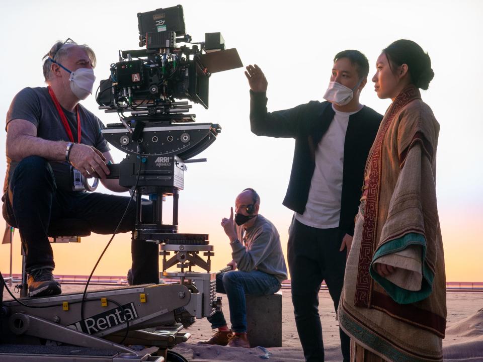 sean savage operating a camera on a platform, with director derek tsang, wearing a face mask, gesturing towards the lens in the front. jess hong, dressed in robes as jin cheng, looks into the camera. they're standing on a sandy set in 3 body problem