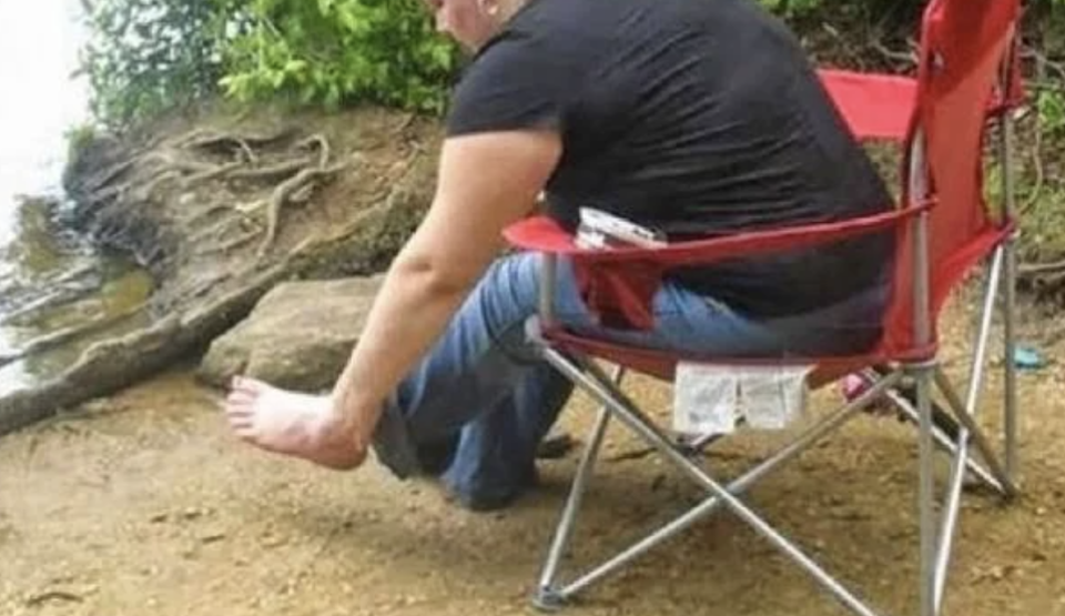 A person sits on a folding chair at the edge of a body of water, dipping their right foot into the water