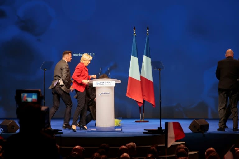 French presidential election candidate for the far-right Front National party Marine Le Pen (2nd L) stands next to her desk on stage as members of the security stop a protester interrupting her speech during a campaign meeting on April 17, 2017