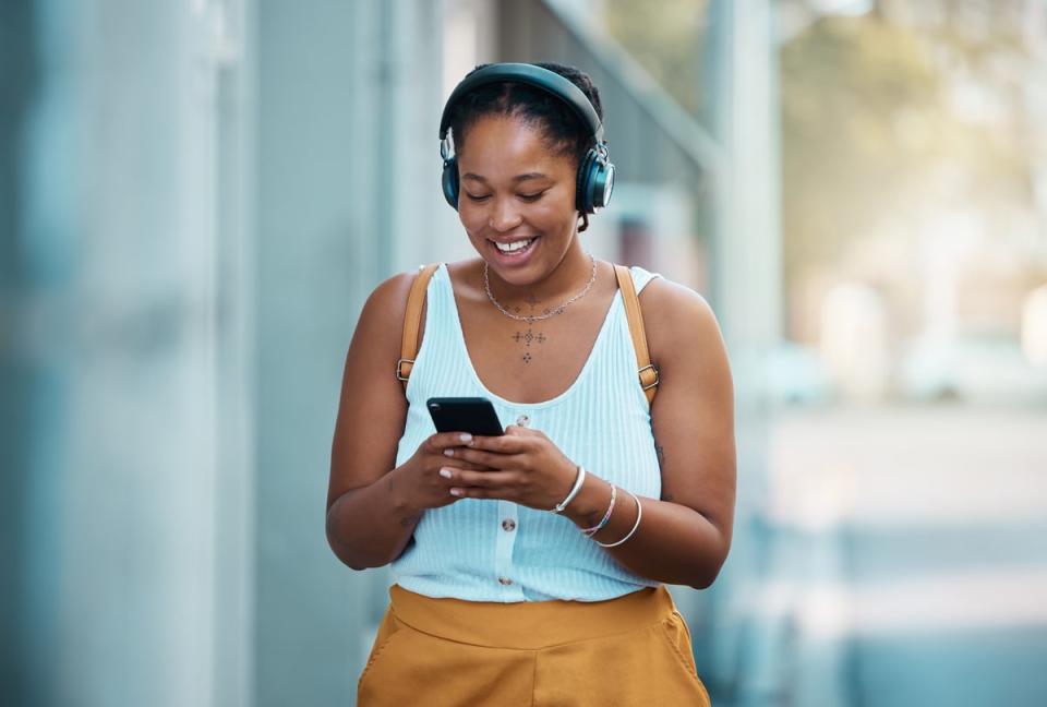 A person wearing headphones smiles while typing on their phone and walking down a street.