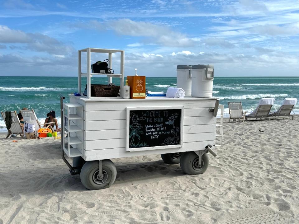 A cart on the beach with sunscreen, water, a towel, and a sign saying "Welcome to the beach."