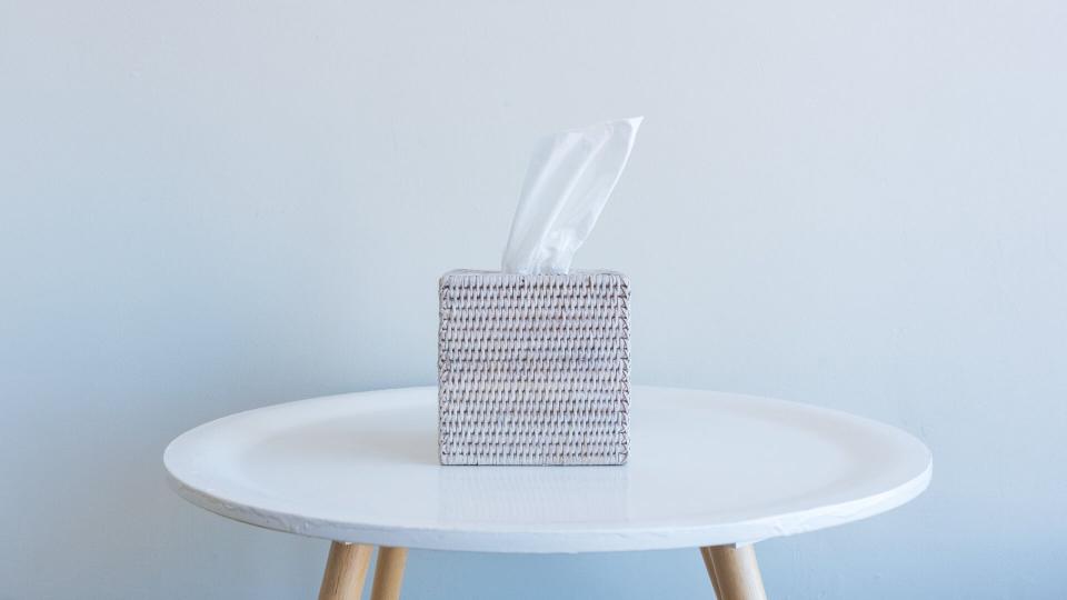 Close up of white tissue in rattan box on small table against neutral wall background - grief concept.