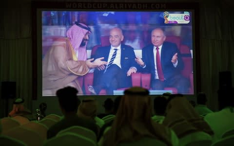 A picture taken on June 14, 2018 in a Saudi football fan tent in the capital Riyadh shows a projector showing the Russia 2018 World Cup Group A football match between Russia and Saudi Arabia, with a clip of Saudi Crown Prince Mohammed bin Salman (L) gesturing as he sits next to FIFA President Gianni Infantino (C) and Russian President Vladimir Putin - Credit: FAYEZ NURELDINE/AFP/Getty Images