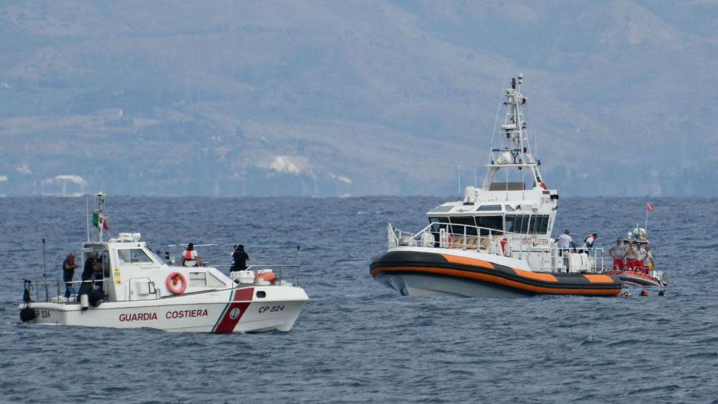 Three white and red Italian coastguard boats in a large stretch of ocean with land in the background. Three divers are in the water next to one of the boats.