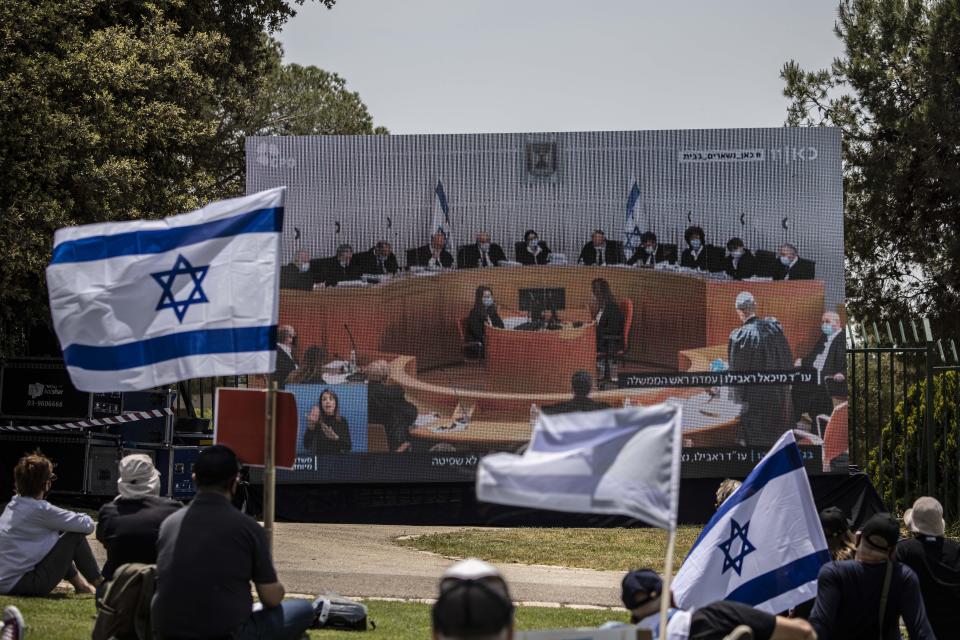 Israeli protesters with hold national flags as they watch a live broadcast of an exceptionally large panel of 11 justices participated in the highly anticipated session in Israel's Supreme Court in Jerusalem, Sunday, May 3, 2020. Israel's high court heard petitions Sunday that seek to block Prime Minister Benjamin Netanyahu from forming a government because he has been charged with serious crimes. (AP Photo/Tsafrir Abayov)
