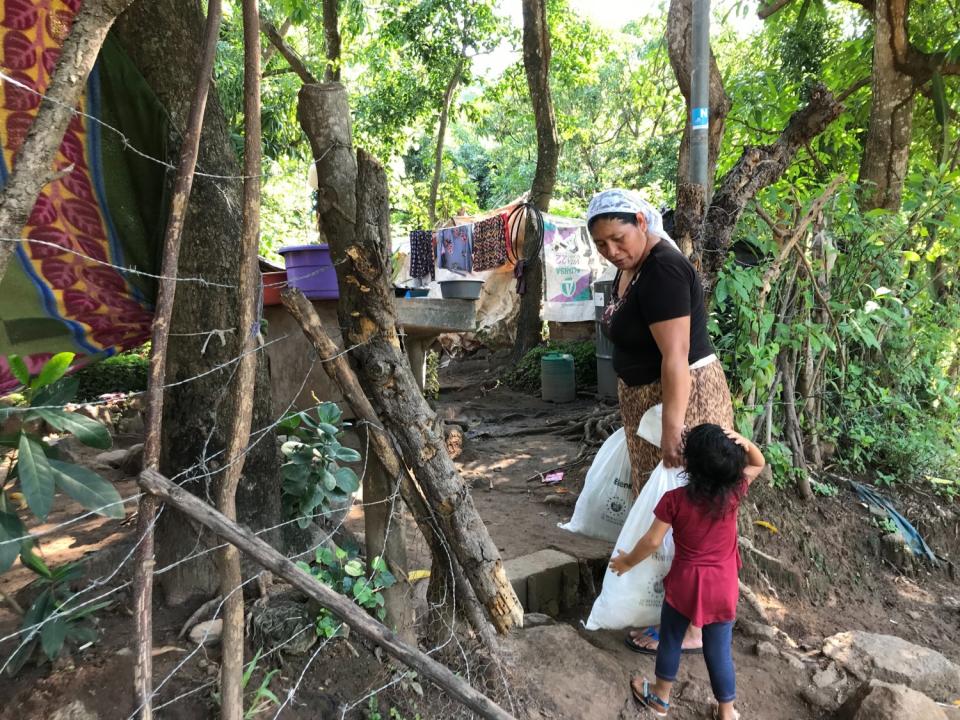 A woman carries packages of food aid while looking down at a child while surrounded by trees.