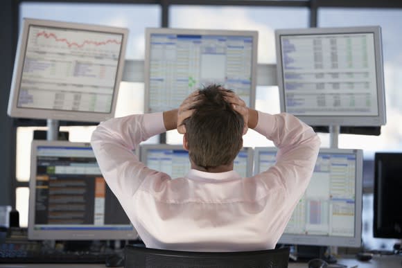 A frustrated stock trader grasping his head while looking at losses on six computer monitors.