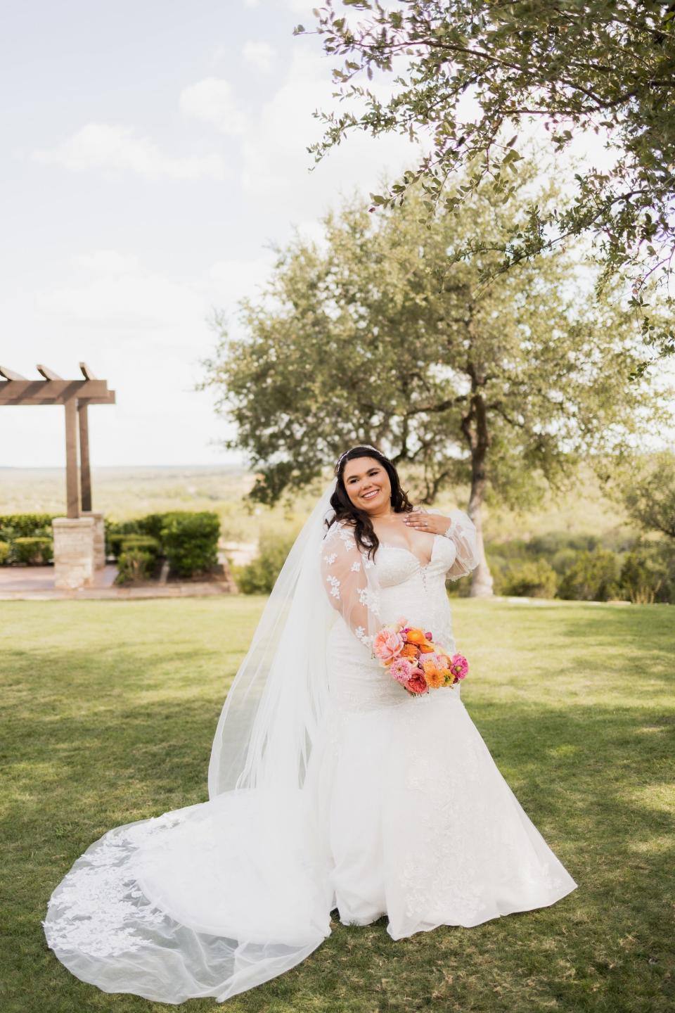 A bride smiles and touches her chest in a field.