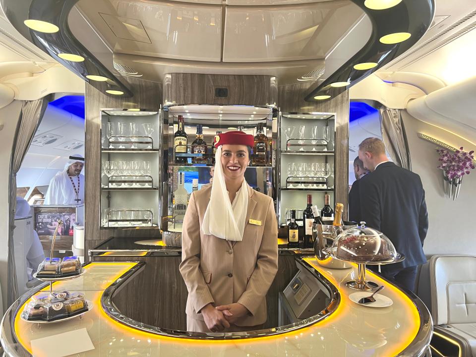 An Emirates flight attendant smiling, stood behind the curved bar onboard an A380 with alcohol and glasses on the shelves and cakes on the bar.