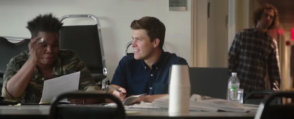 Kyle Mooney suspiciously looking in on Colin Jost talking to Leslie Jones