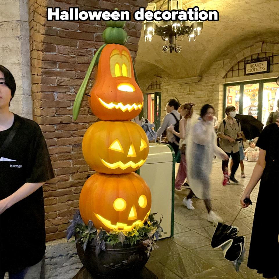 People walk past a tall pumpkin stack decoration shaped like cartoon characters with jack-o'-lantern faces, under an archway with chandeliers