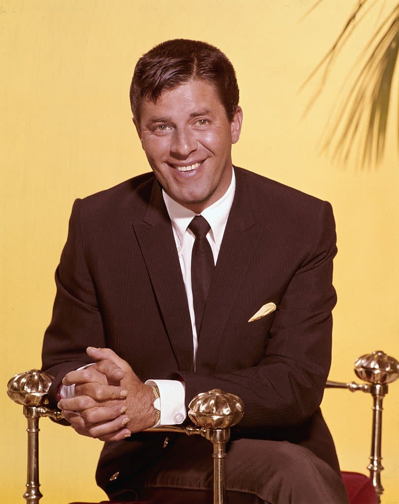 Jerry Lewis smiling while sitting, wearing a dark suit and tie with a light-colored pocket square. He is against a plain background with a hint of foliage