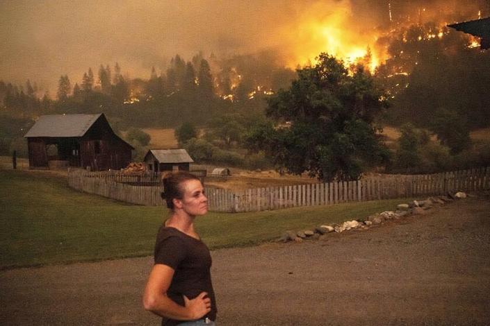 A woman stands near a house as wildfire burns a nearby ridge.