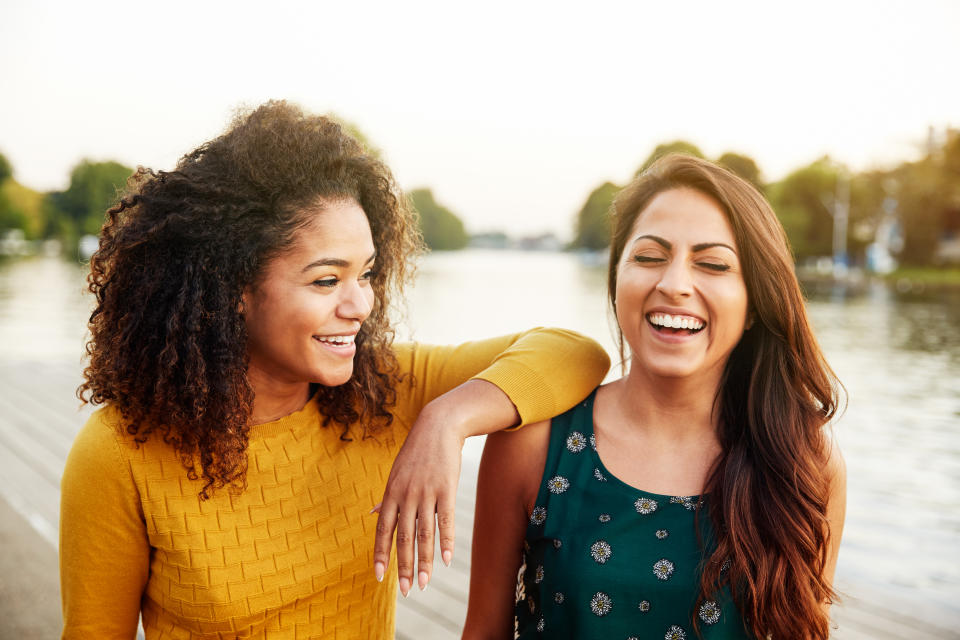 Two women, one with an afro hairstyle and wearing a yellow long-sleeved top, and another with long brown hair, wearing a sleeveless green top, laugh together