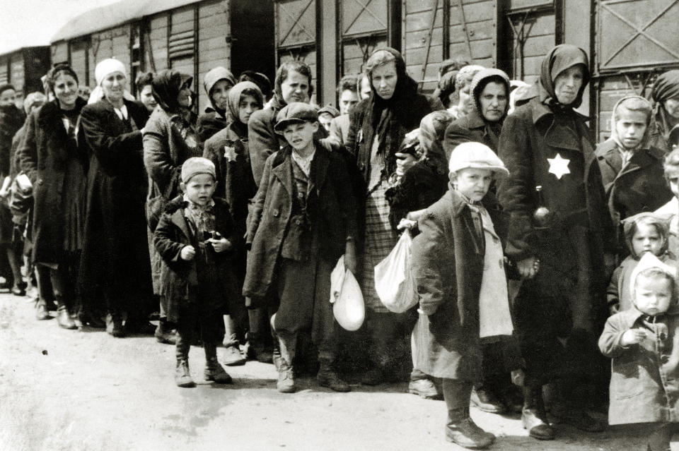 A long line of women and children, some wearing large Stars of David, forms beside a railroad cattle transport car.