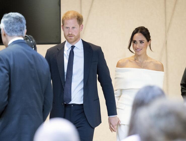 Prince Harry in a suit and Meghan in a white dress hold hands as they walk through a crowd