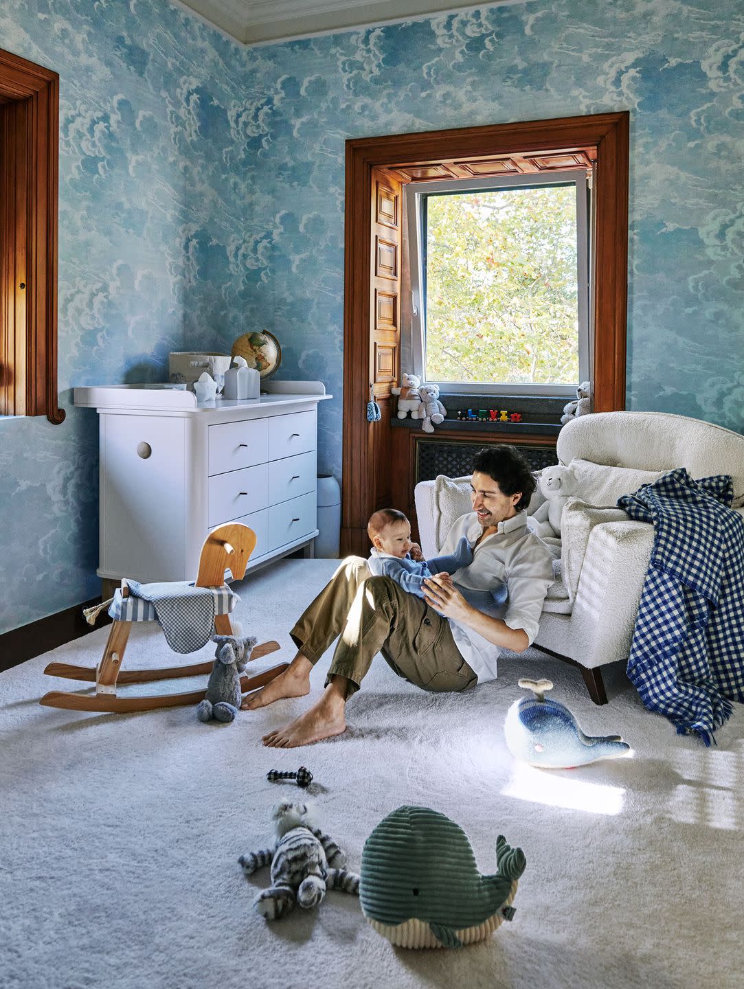 a man and his baby sit on the floor of a nursery with cloudlike pattern wallpaper, two windows with carved woodworking, a boucle chair, a white chest of drawers, a wood rocking horse, and stuffed toys