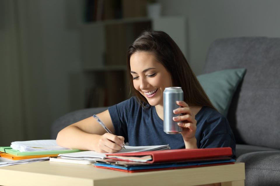 A person writes notes in a notebook while consuming an energy drink.