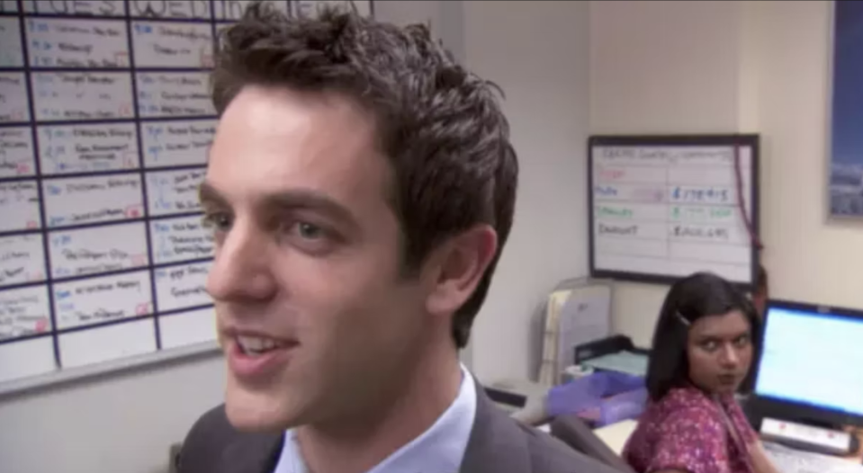 Man in suit in office setting with woman in background; scene likely from TV show "The Office"