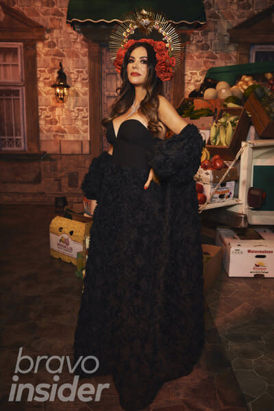 Adriana De Moura wears a black, off shoulder, gown and a rose-adorned headpiece, in front of a Mexico City inspired set.