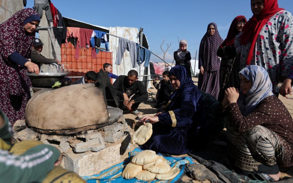 People wait while a woman prepares food, as displaced Palestinians, who fled their houses due to Israeli strike, shelter in a camp in Rafah