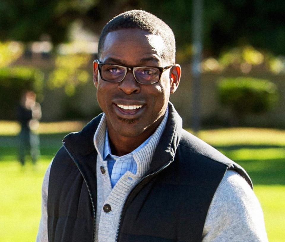 A man in glasses smiles wearing a layered sweater and vest, standing outside