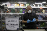 Liquor store worker Mario Valle puts on protective gloves behind a sign requiring face masks in the Vermont Square neighborhood of Los Angeles, Thursday, May 21, 2020. While most of California is welcoming a slight return toward normal this holiday weekend, Los Angeles will not be joining the party. The nation's largest county is not planning to reopen more widely until the next summer holiday, July 4th, because of a disproportionately large share of the state's coronavirus cases and deaths that have hampered the county's ability to rebound and meet strict criteria to get more people back to work.(AP Photo/Jae C. Hong)