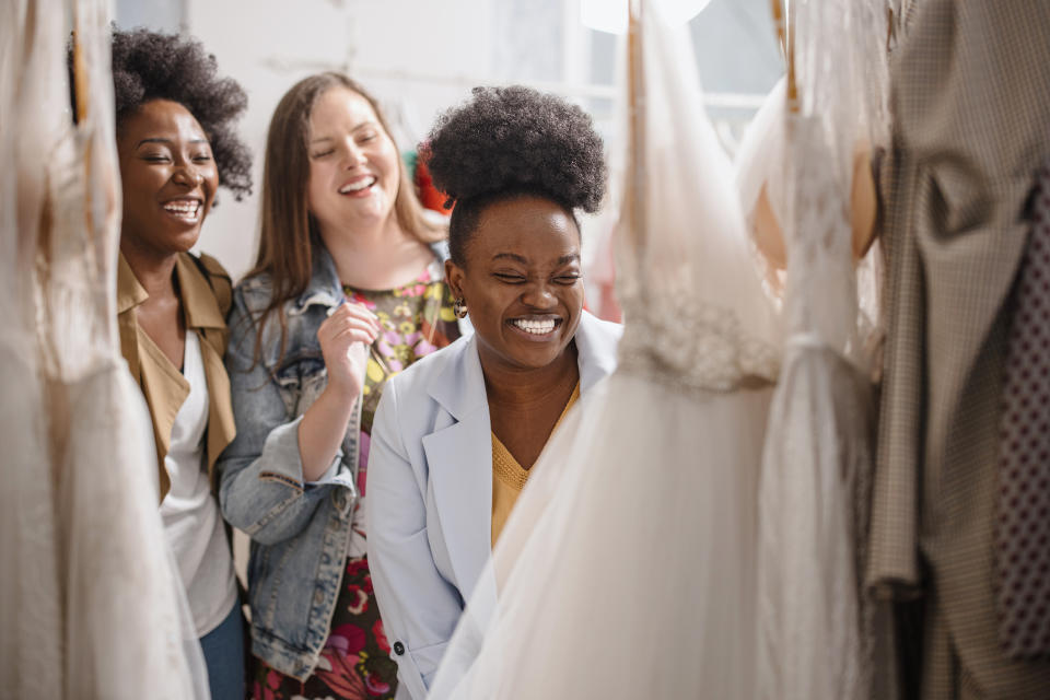 Three women happily browse wedding dresses in a boutique. One wears a light blazer, another a floral dress, and the third a jacket, all smiling and enjoying the moment