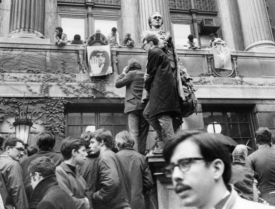 FILE - Part of some estimated 300 students at Columbia University are shown milling around Hamilton Hall on the campus in New York, April 24, 1968. The students are protesting the construction of a gymnasium in a public park and the university's participation in a defense-related program. A couple of students stand on pedestal of the statue of Alexander Hamilton while others hang a poster of Stokely Carmichael from the balcony of the building along with a Viet Cong flag. (AP Photo/Jacob Harris, File)