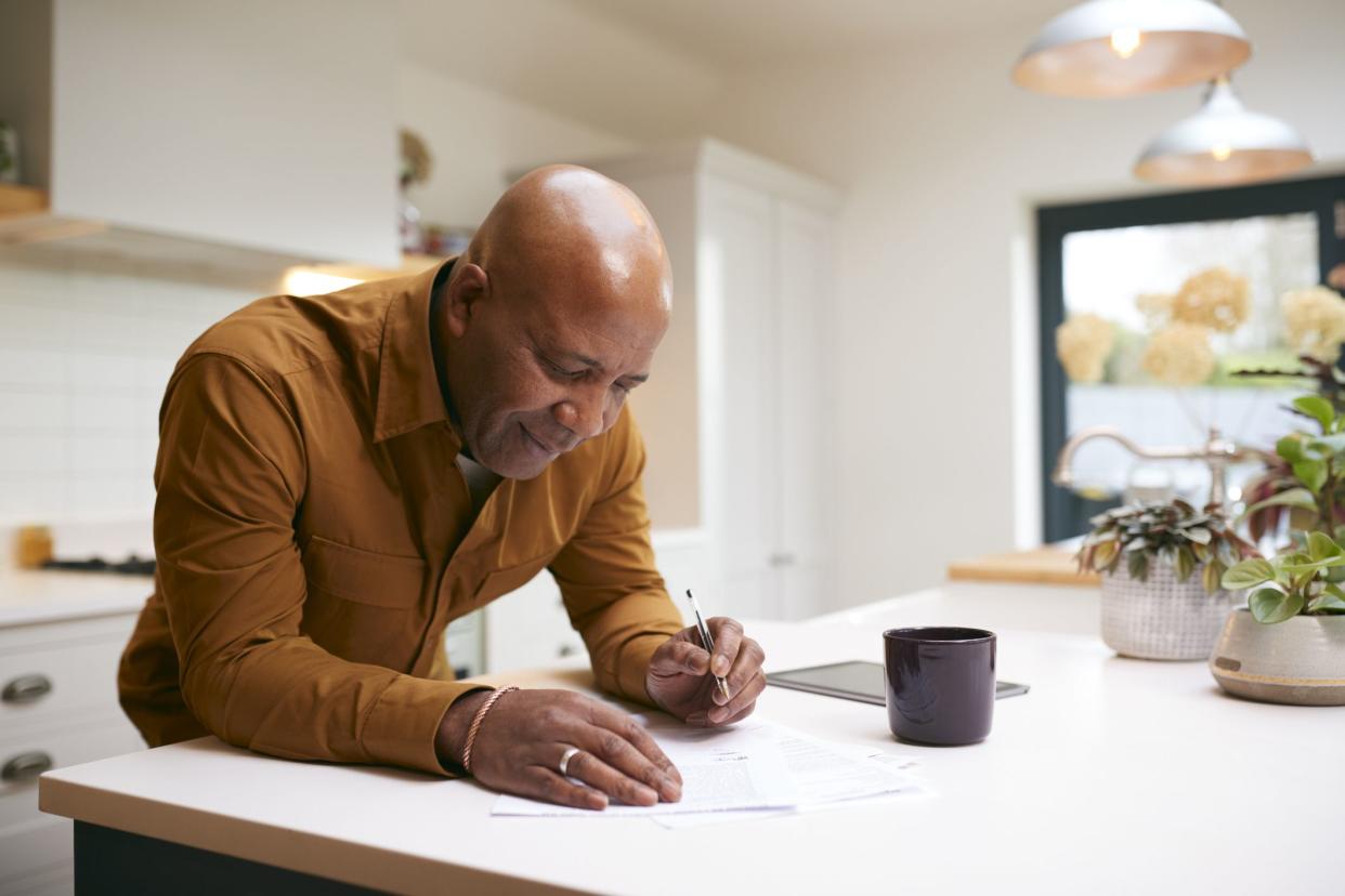 Mature Man Reviewing And Signing Domestic Finances And Investment Paperwork In Kitchen At Home