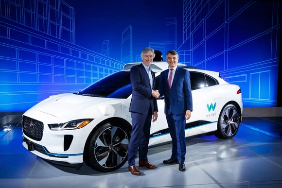 The two executives are standing in front of a white Jaguar I-Pace SUV with Waymo logos and visible self-driving sensor hardware.