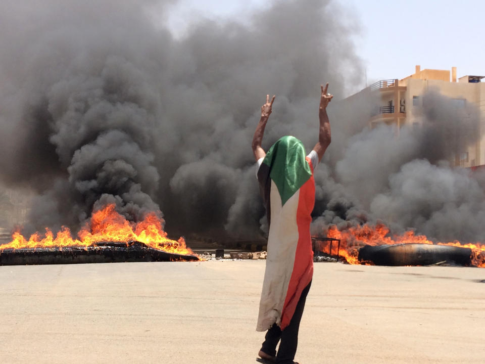 A protester wearing a Sudanese flag flashes the victory sign in front of burning tires and debris on road 60, near Khartoum's army headquarters, in Khartoum, Sudan, Monday, June 3, 2019. Sudanese protest leaders say at least 13 people have been killed Monday in the military's assault on the sit-in outside the military headquarters in the capital, Khartoum. The protesters have announced they are suspending talks with the military regarding the creation of a transitional government. (AP Photo)