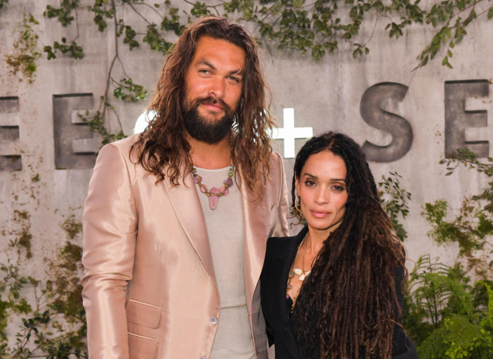 Jason Momoa in a satin suit and Lisa Bonet in a black outfit at an event, standing together in front of a backdrop with greenery and text