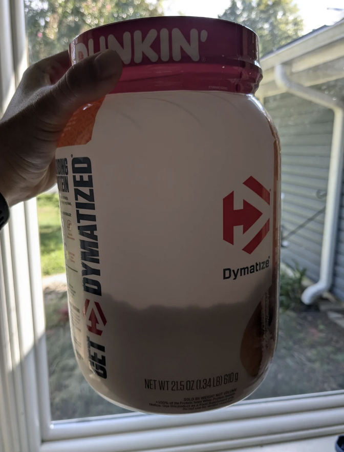 A hand holds a large jar of Dymatize Dunkin' protein powder by a window in natural light