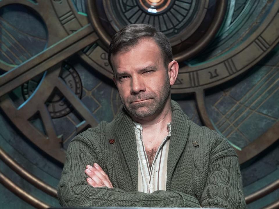 An on-set photo of Critical Role's Liam O'Brien. He's wearing a dark green Cardigan and a striped, button-down shirt.
