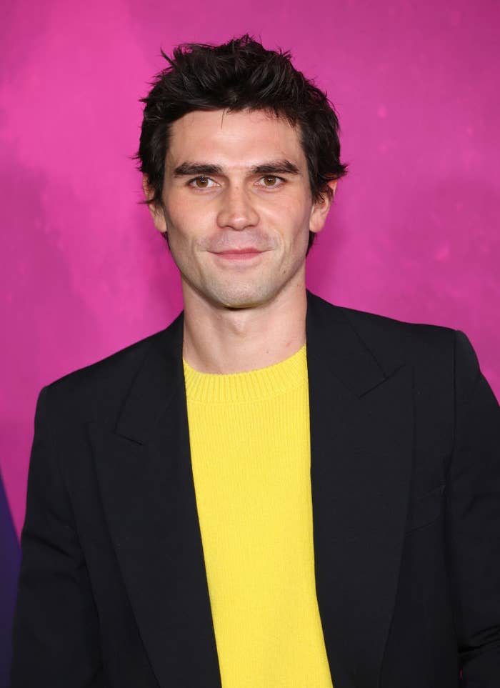 A man is seen from the chest up, wearing a black suit jacket over a yellow sweater, with a pink backdrop behind him