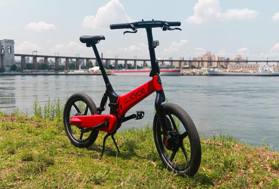 A red Gocycle G4i+ electric folding bike on grass, with water and blue skies in the background.