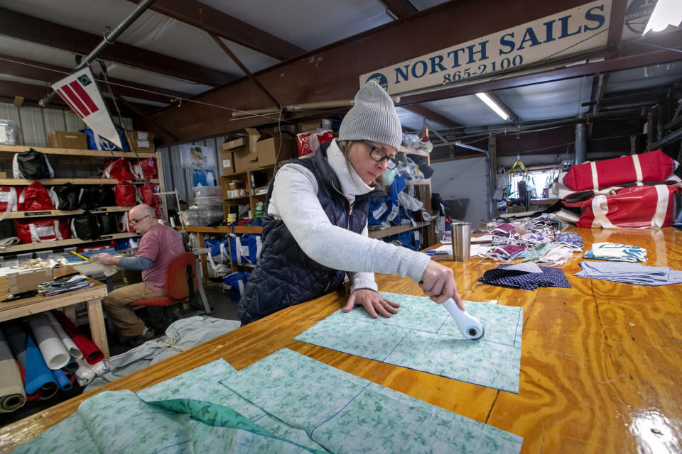 In this Monday, March 23, 2020, photo, Karen Haley cuts cotton fabric for masks to be given to caregivers during the coronavirus outbreak, at the North Sails shop in Freeport, Maine. The sail-maintenance business has converted part of its operation towards stitching masks instead of sails. Owner Eric Baldwin stitches masks in background. (AP Photo/Robert F. Bukaty)
