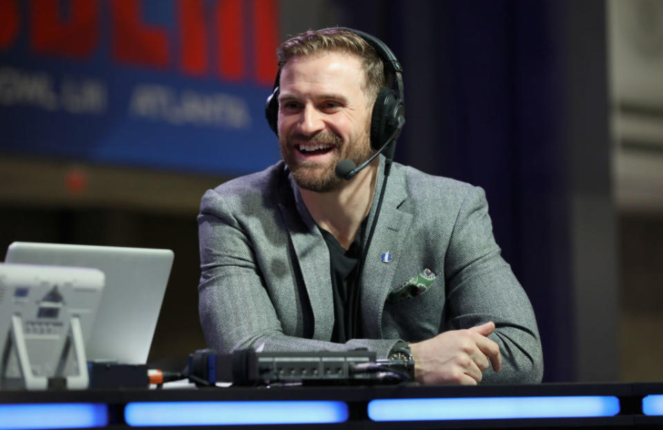 Chris Long, wearing a headset and gray blazer, smiles while seated behind a broadcast desk