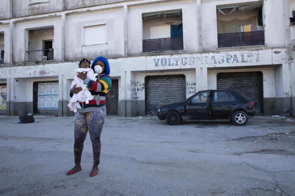 In this photo taken on Monday, April 27, 2020, a woman wearing a sanitary mask to protect against COVID-19 holds her daughter as she walks past a building in Castel Volturno, near Naples, Southern Italy. They are known as “the invisibles,” the undocumented African migrants who, even before the coronavirus outbreak plunged Italy into crisis, barely scraped by as day laborers, prostitutes and seasonal farm hands. Locked down for two months in their overcrowded apartments, their hand-to-mouth existence has grown even more precarious with no work, no food and no hope. (AP Photo/Alessandra Tarantino)