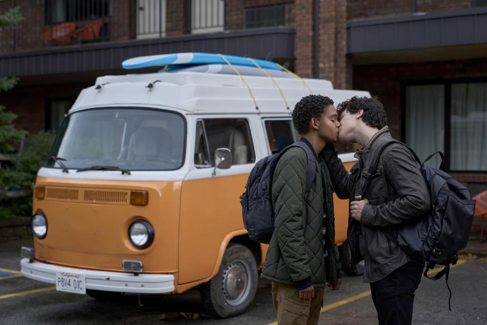 Two boys kiss in front of an orange van with a surfboard on top. They are both wearing jackets and backpacks