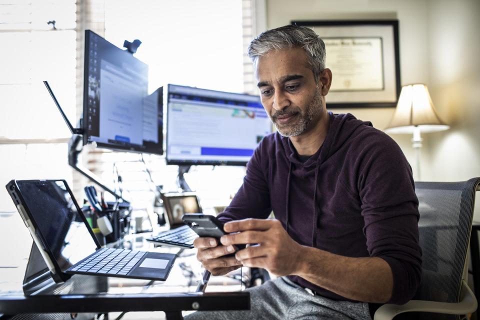 A man at a desk with several computer screens and looking at his cellphone.