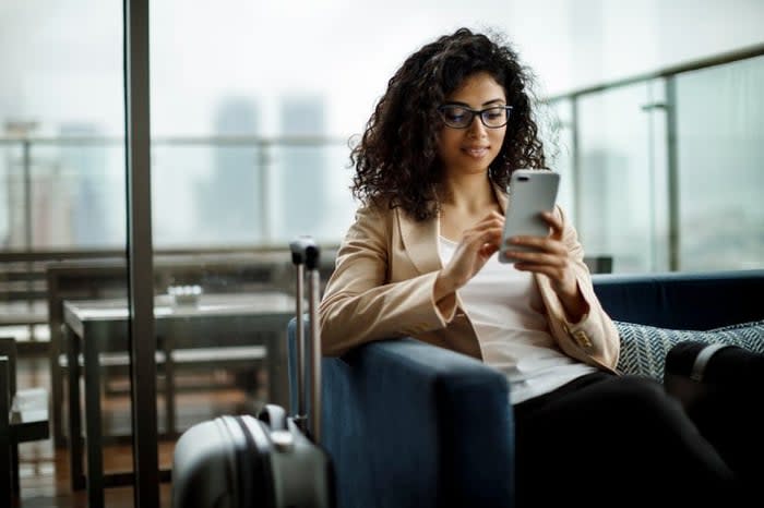 A woman sitting in an airport lounge next to her suitcase and looking at her phone.