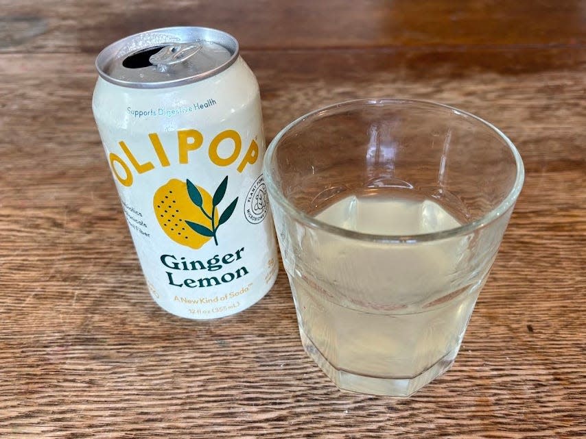 An open can of ginger-lemon Olipop next to a small, clear glass with pale-yellow liquid inside. Both are sitting on a wooden table.