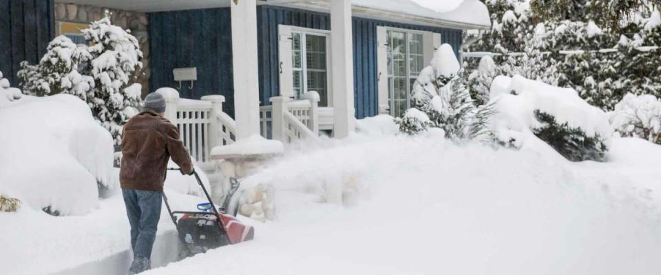 Man using snowblower to clear deep snow on driveway near residential house after heavy snowfall