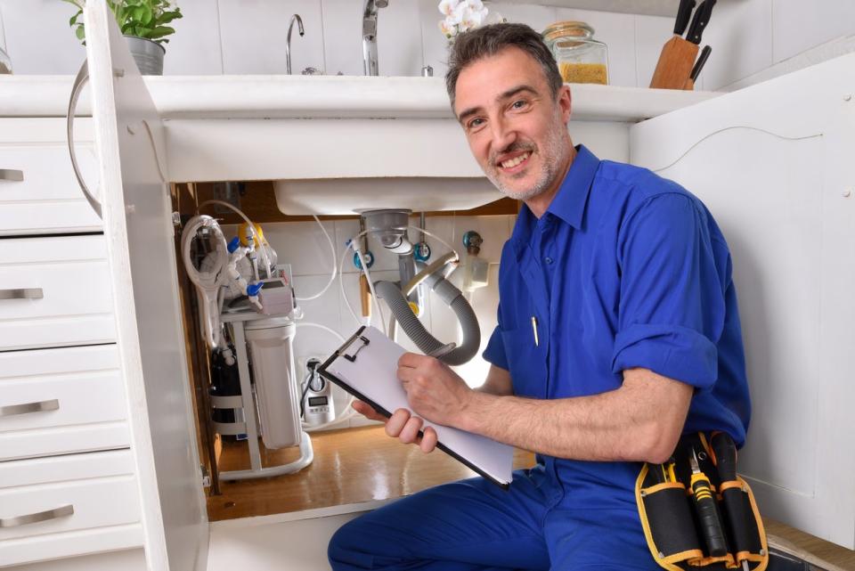 A man smiles while checking items off a list on a clipboard while inspecting plumbing under a sink.